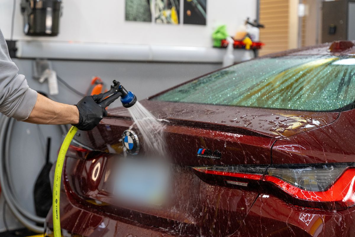 A technician rinsing the trunk of a red BMW M4, water beading on the rear glass and trunk lid