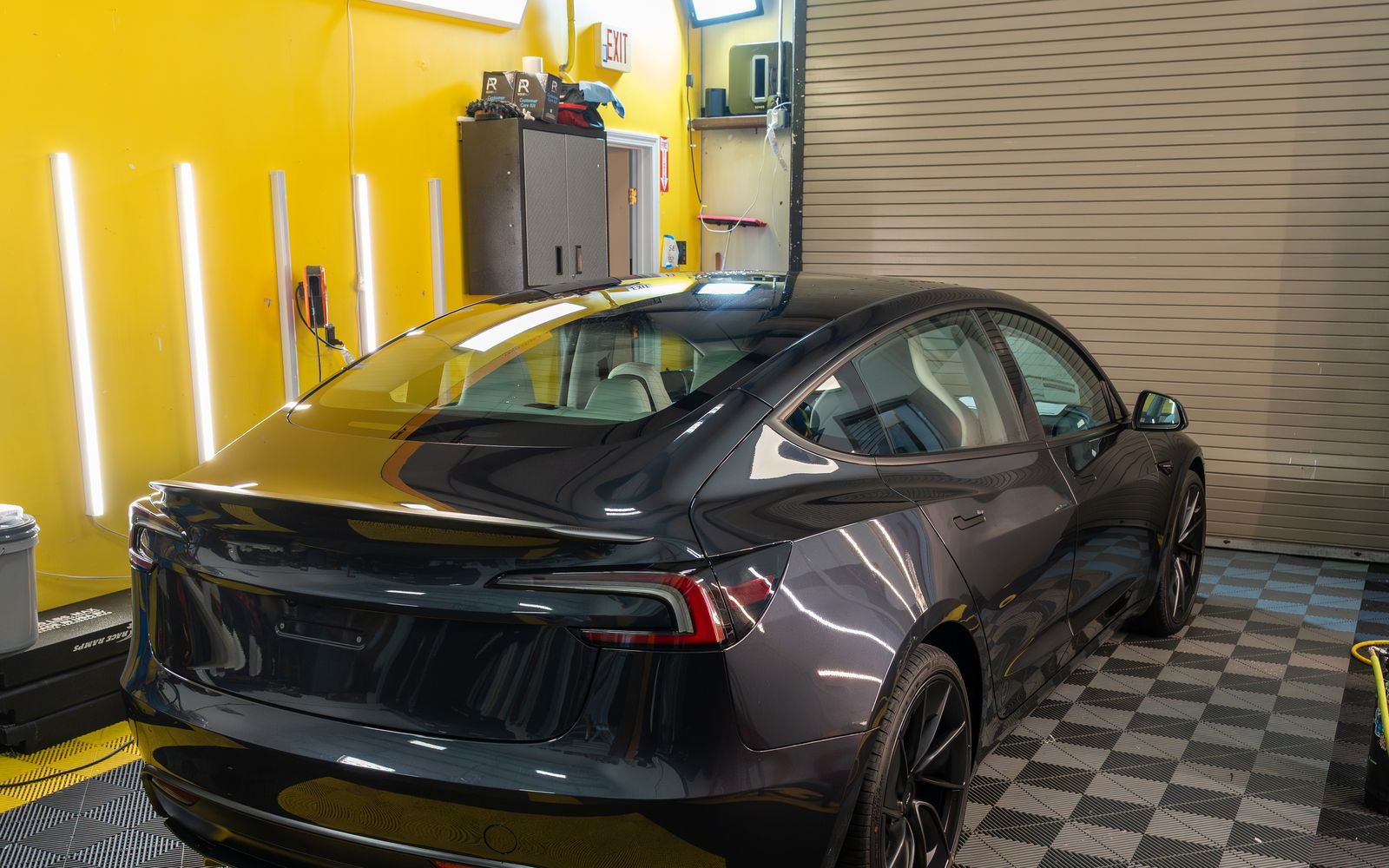 A grey Tesla Model 3 from the rear three quarter in the ACP bay, against a yellow wall