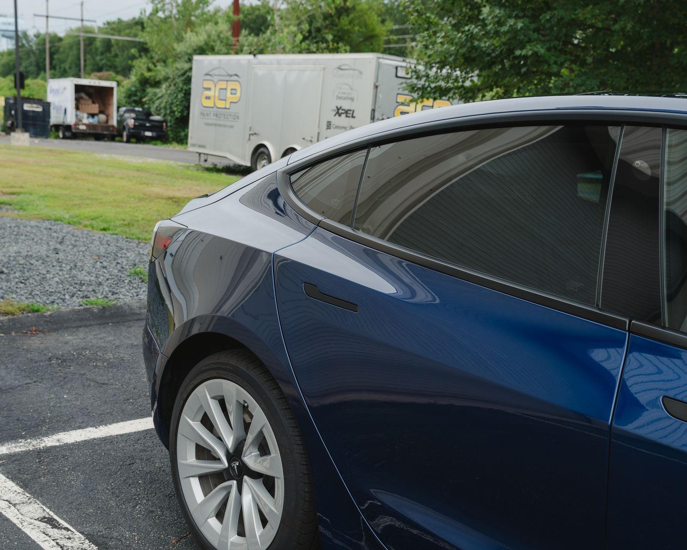 The dark tinted rear glass of a blue Tesla Model 3, with ACP's trailer parked behind it